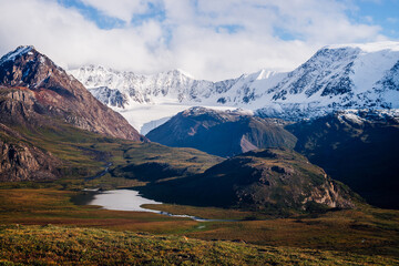 Awesome picturesque view to mountain range in sunny day. Beautiful green highland relief with glacier, lake, river, white snowy tops and low clouds in sunlight. Scenic landscape with snowy mount ridge