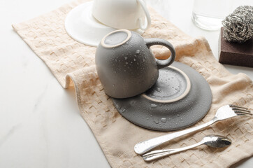 Clean cups and saucers,fork and spoon drying on the kitchen towel.Brown sponge, dish scraper, dishes on the white table.Empty space