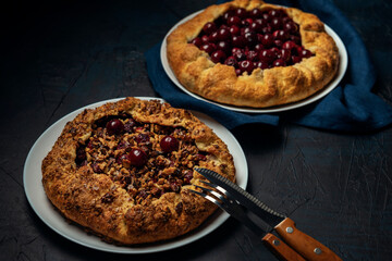 Two galettes with ripe red cherry and walnut filling on dark blue background. Homemade sweet open pie with napkin and cutlery.