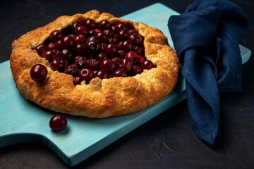 Galette with ripe red cherry filling with napkin on blue cutting board. Homemade sweet open pie on dark background.