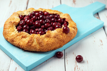 Galette with ripe red cherry filling with napkin on blue cutting board. Homemade sweet open pie on white wooden background.