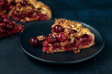 A piece of galette with ripe red cherry filling on black plate. Homemade sweet open pie on dark textured backdrop.