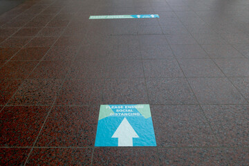 A floor sign with arrow says 'please ensure social distancing' on a tiled floor with further sign in distance.Image
