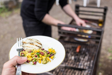 Medium wide view of left hand holding fork with thumb, plate with grilled halloumi cheese, yellow rice with beans, parsley and Mexican nopales. Person in black prepares food next to live grill