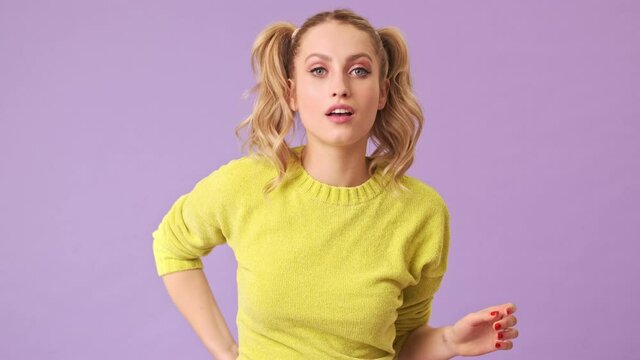A charming blonde in a yellow sweater prepares for a date, straightens her ponytails on her head, takes out a makeup brush and begins to paint her face in an isolated studio on a purple background