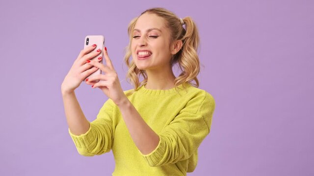 A beautiful blonde takes a selfie on the phone and grimaces in an isolated studio on a purple background
