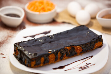 rectangular carrot cake, covered with 70% cocoa chocolate, with unfocused ingredients in the background..