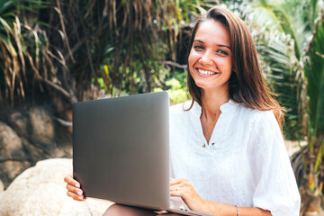 Young girl, business lady working on a laptop while traveling sitting among tropical palm trees on the stones