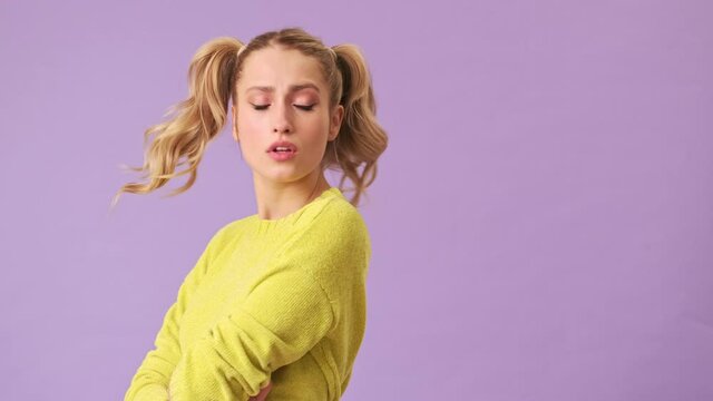 A cute blonde in a yellow sweater is unhappy and upset by the behavior of someone, sadly turned in the opposite direction, looking at the floor in an isolated studio on a purple background