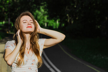Naklejka premium Portrait young woman in white top touching her hair and enjoying sun light on the drive way in the park . Solo outdoor activity. End quarantine. Alone teenage girl in the nature. Close up