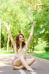 Girl in pink shorts and white top smiling and sitting in the middle of the road in the park with legs crossed and hands up in victory sign. Green grass and trees around. End of pandemia and quarantine