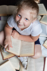 A smiling little girl is sitting on the floor with books. Education and training. Black background. Vertical. Top view.