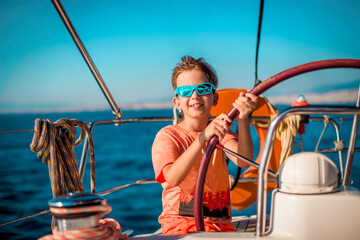 A handsome boy of 9-10 years old at the helm of a yacht in bright orange clothes, blue glasses...