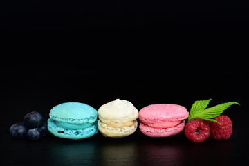 Three sweet macarons, in the colors of the French flag, lie side by side against a dark background, with blueberries and raspberries