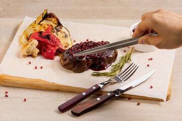 Steak with lingonberry sauce with grilled vegetables are on the wooden background and hand holds a tweezer. Processes of food photo.