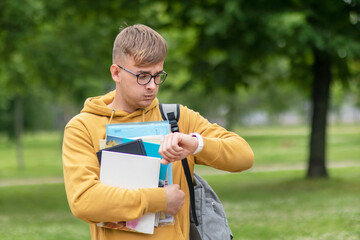 Stressed anxious young guy is late, busy university or college student or pupil with books, textbooks in glasses looking at his wrist watch, checking time in a hurry, rushing to lessons, exam. No time