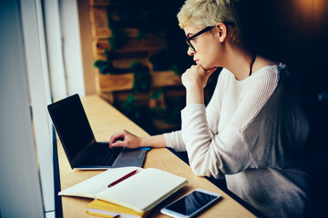 Pensive creative manager in eyeglasses reading news on laptop analyzing information for report,...
