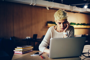 Beautiful hipster girl with trendy short hairstyle watching movie on laptop during leisure in...