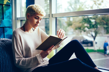 Attractive trendy dressed hipster girl reading interesting book resting in coworking space sitting...