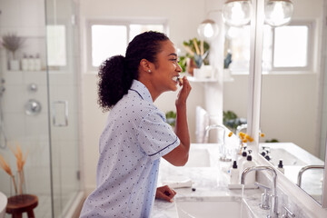 Woman Wearing Pyjamas Standing At Sink Brushing Teeth In Bathroom