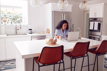 Woman Wearing Pyjamas Sitting In Kitchen Working From Home On Laptop