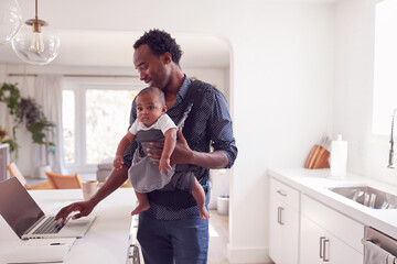 Father With Baby Daughter In Sling Multi-tasking Working From Home On Laptop