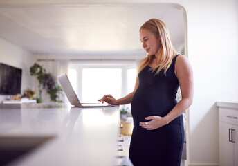Fototapeta na wymiar Pregnant Woman Standing By Kitchen Counter Working From Home On Laptop