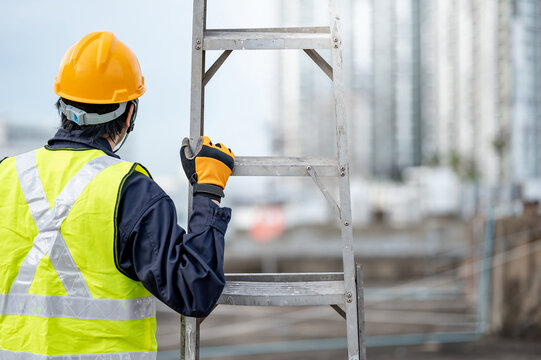 Asian maintenance worker man with safety helmet and green vest carrying aluminium step ladder at construction site. Civil engineering, Architecture builder and building service concepts