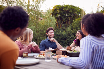 Multi-Cultural Friends At Home Sitting At Table Enjoying Food At Summer Garden Party