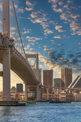Fototapeta premium Sunset sky on the suspension Rainbow Bridge in the Tokyo Bay with barge towed by a tugboat sailing in Tokyo port and the skyscrapers of the Shibaura district in the background.