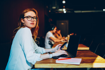 Serious businesswoman in eyewear synchronizing multimedia from smartphone storage to laptop,...
