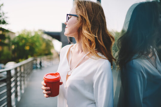 Dreamy young woman holding coffee to go cup recreation during leisure outdoors during spring day,pensive hipster girl looking away standing near publicity area feeling relax on morning street