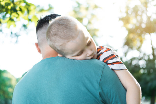 A Young Child Is Sleeping In His Father's Safe And Protective Embrace While On Walk. Portrait Of Cute Adorable Blond Caucasian Toddler Boy Sleeping On Fathers Shoulder Outdoors. Single Father Concept.