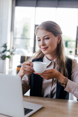 selective focus of happy businesswoman holding cup of coffee and looking at laptop