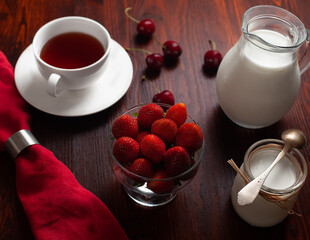 Healthy breakfast with strawberries, sweet cherries, yoghurt, tea and teapot. Served with red napkin. Dark background.