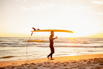 Young bearded surfer in wetsuit with yellow surfing longboard walk from water at sunset ocean....