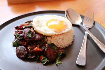 Chinese sausage fried with basil and fried egg with rice in the black round dish, with spoon and fork.