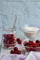 Currants and cherries in a glass jar, a glass plate with berries and sugar on the background of a table of white boards; light background for summer recipes for making homemade jam