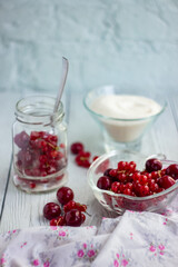 Currants and cherries in a glass jar, a glass plate with berries and sugar on the background of a table of white boards; light background for summer recipes for making homemade jam