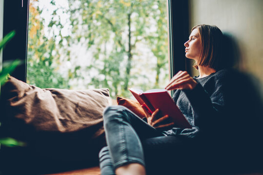 Thoughtful young woman looking out of window pondering on idea and plot of new bestseller of favourite author resting on comfortable windowsill in stylish coworking space.Copy space area