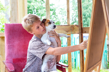 A blond-haired boy sits in a pink chair and holds the dog Jack Russell Terrier in his arms.Caucasian teenager draws on an easel, plener.Painting outdoors, not in studio.Portrait of a child with a dog