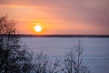 Landscape of sundown over a frozen river and snowy forest in the background.