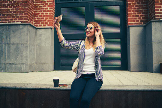 Happy attractive female blogger in stylish eyeglasses posing while making selfie on smartphone camera for updating profile picture.Positive good looking student taking photo during free time outside