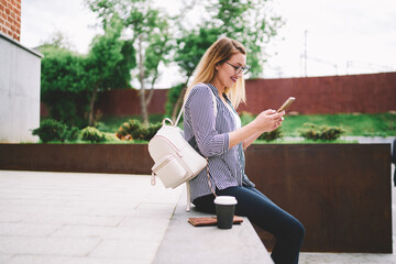 Cheerful hipster girl in spectacles outdoors check mail by new cellular phone with 4g connection, fashionable positive woman with trendy backpack sending text messages using modern mobile phone