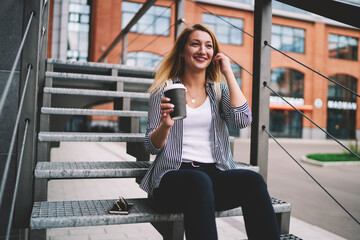 Cheerful successful hipster girl 20 years old sitting on stairs with takeaway coffee in hand, positive female teenager with smile on face relaxing outdoors and waiting friend on urban setting