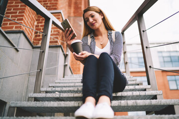 Smiling female student 20 years old watching new episode from serial via modern telephone while resting outdoors, happy hipster girl sitting at stairs enjoying coffee and messaging via mobile phone