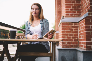 Positive hipster girl writing happy moments of day in textbook while using telephone, carefree female student looking away while creating article for blog in notebook and resting at street cafe