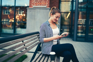 Gorgeous positive female blogger sharing new ideas for content in personal website with followers during walk at urban setting.Happy cute woman resting outdoors with modern device and tasty coffee