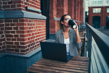 Young creative female freelancer in spectacles enjoying aroma coffee while sitting at wooden table with laptop computer.Talented student drinking tasty beverage during distantly work at street