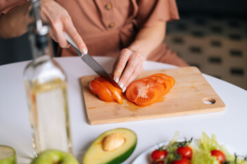 Close-up of hand of woman cutting fresh tomato using kitchen knife on wooden cutting board. Young woman cutting fresh organic tomato with a knife for vegetable salad.
